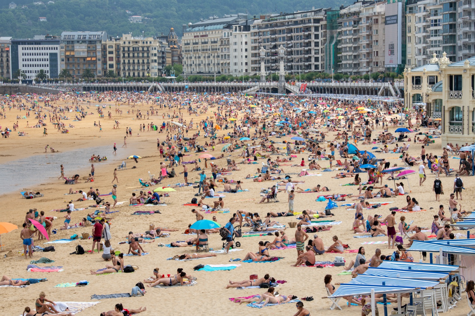 A city beach packed edge to edge with umbrellas and swimmers, belle epoque buildings rising behind the promenade
