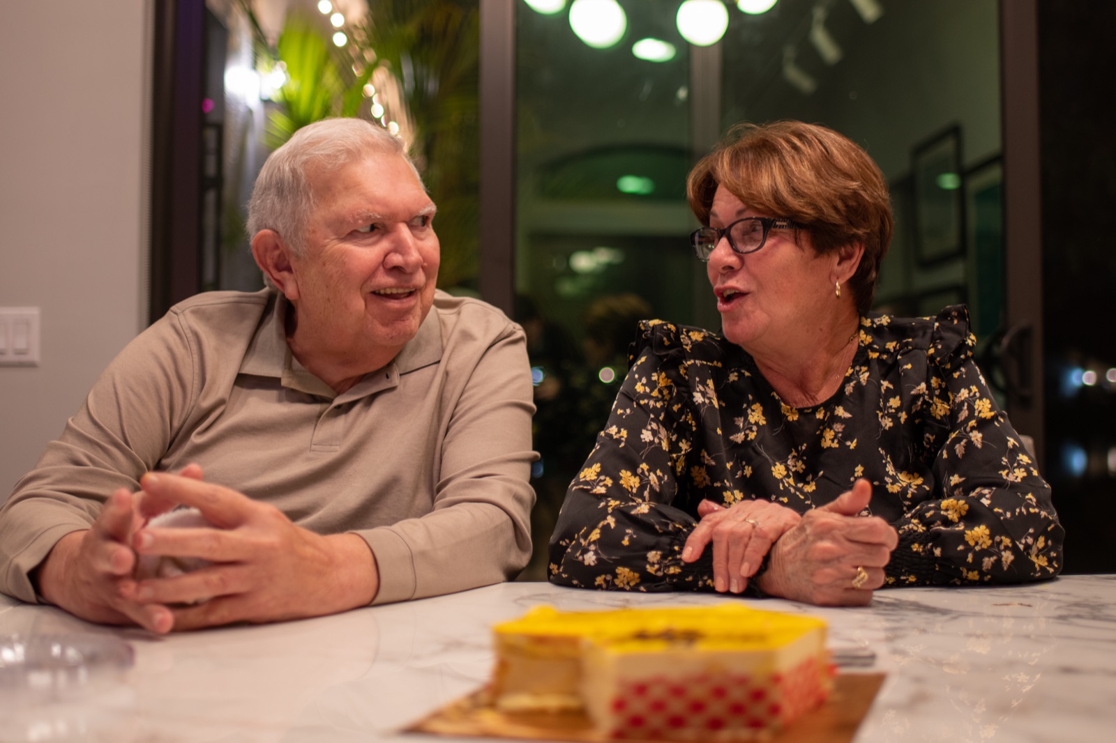 My parents at a marble counter, caught mid-laugh looking at each other, a yellow cake waiting in the foreground